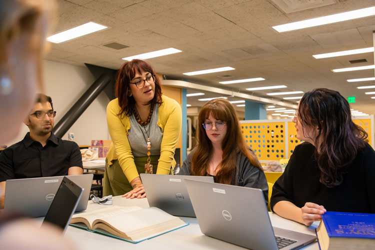 An instructor stands near a table with a group of students seated with laptops