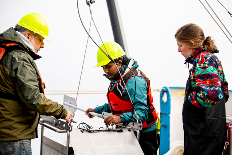 Three people on a industrial boat deck looking at charts, two of them wearing hardhats