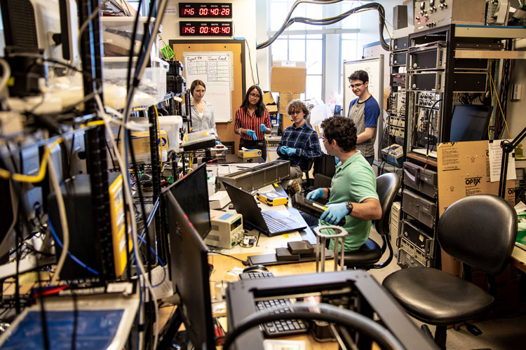 group of five young undergrad researchers talk to each other in room with electronic equipment lining the walls and desks 