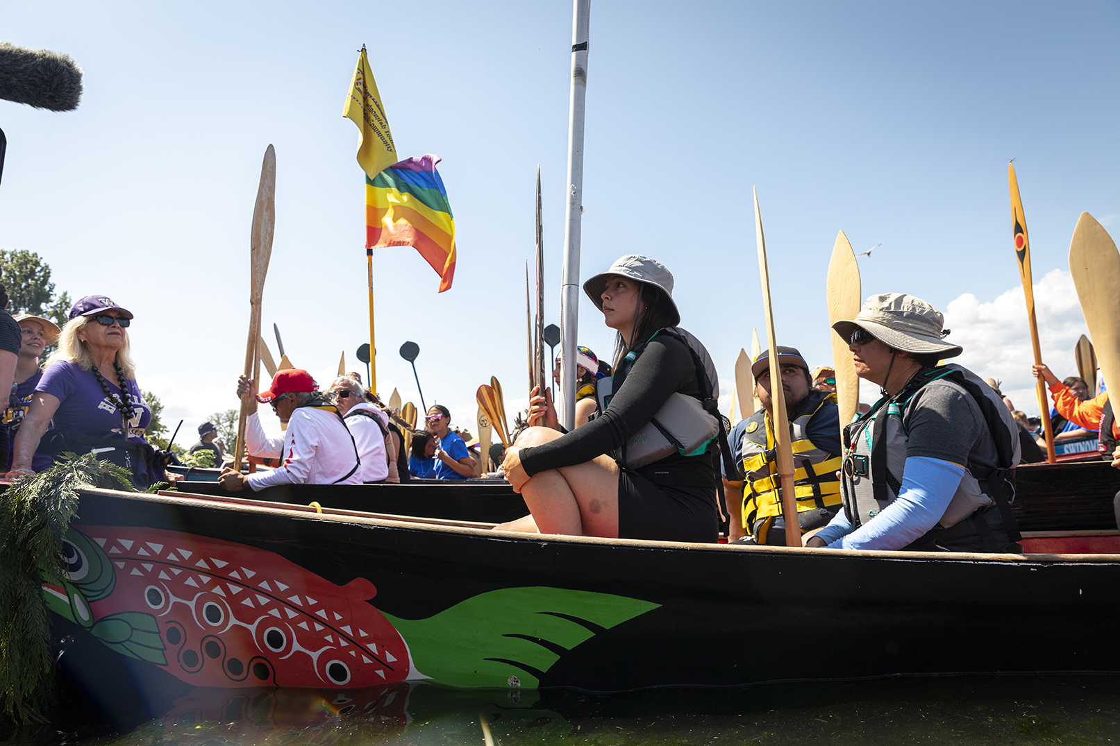 Woman seated in painted tribal canoe with UW flag and people in the background also in canoes with other tribal flags