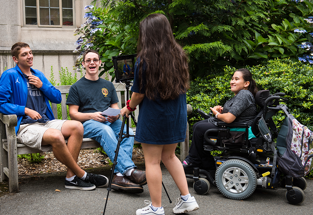 Two students sit on a bench while another person sets up a video camera to film them and a person in a motorized wheel chair talks to camera operator