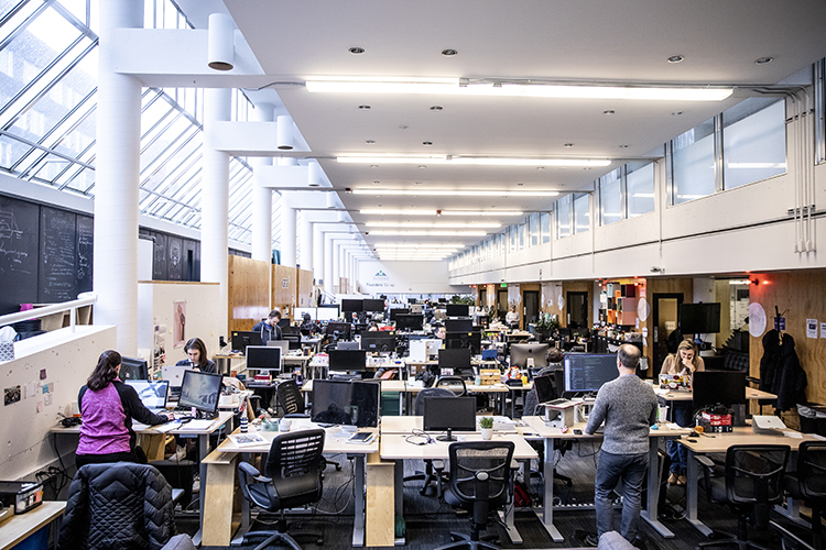 View of a long corridor of open desks with monitors full of people