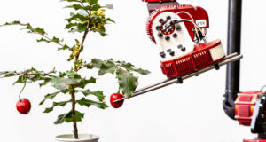 a large, silver and red colored robotic arm gently picks a cherry tomato from a plant using a pair of thin chopsticks