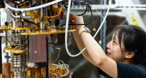 A woman scientist wearing goggles is using tools to adjust pieces on a large cylindrical machine with tubes and hoses