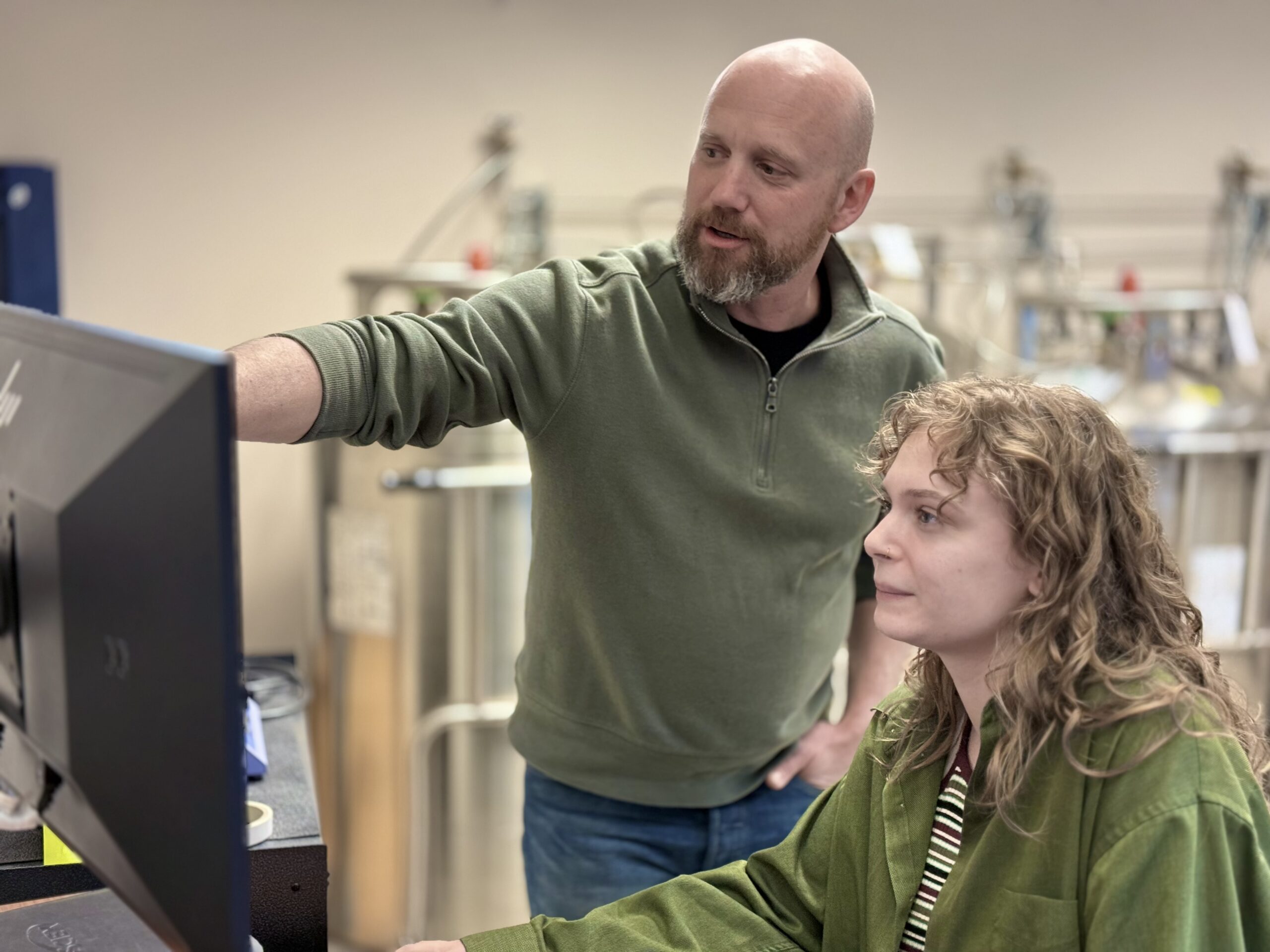 Younger woman student sitting in front of computer monitor with Man standing beside her pointing at monitor
