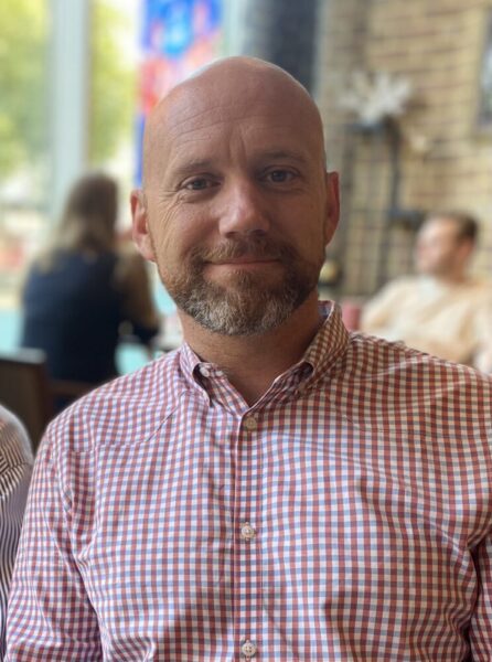 Dale Whittington facing the camera smiling, bald with a light brown beard, wearing a red and white checkered shirt